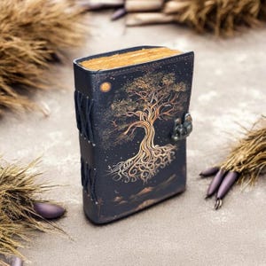 May include: A black leather journal with a gold tree design and a metal clasp. The journal is closed and has a rustic look.