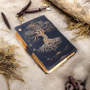 May include: A black leather journal with a gold tree design on the cover. The tree has intricate roots and branches, and the background is a dark blue with gold stars. The journal is closed and lying on a brown surface.