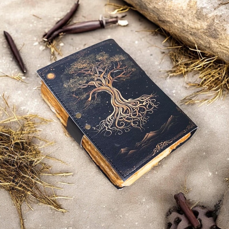 May include: A black leather journal with a gold tree design on the cover. The tree has intricate roots and branches, and the background is a dark blue with gold stars. The journal is closed and lying on a brown surface.
