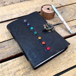 May include: Black leather journal with a silver clasp and a pen. The cover features a circular design with seven colorful gemstones.