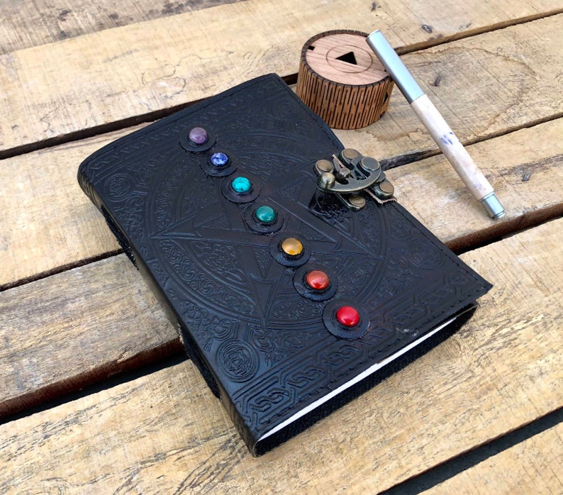 May include: Black leather journal with a silver clasp and a pen. The cover features a circular design with seven colorful gemstones.
