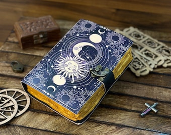 Puede incluir: Un diario de cuero azul oscuro con un diseño celestial que presenta un sol, una luna y estrellas. El diario tiene un cierre de latón y está cerrado.