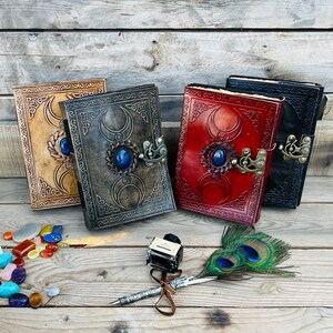 Op de afbeelding: Drie lederen dagboeken met ingewikkelde ontwerpen en edelsteenaccenten. Elk dagboek heeft een drievoudig maansymbool en een blauwe edelsteen. De dagboeken zijn in een rij op een houten oppervlak gerangschikt met een pauwenveer en een inktpot.
