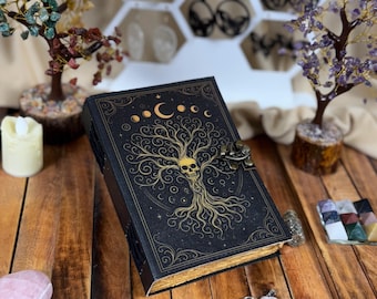 May include: A black journal with a gold tree of life design, a skull, and crescent moons. The journal has a metal clasp and gold-edged pages. The cover is embossed with a celestial design. The journal is on a wooden surface.