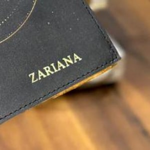 May include: Close-up of a black leather wallet with gold lettering that reads "ZARIARA".