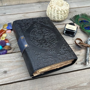Op de afbeelding: Een zwart lederen gebonden dagboek met ingewikkelde gegraveerde bloemmotieven en een blauwe leren band. Het dagboek is gesloten en ligt op een houten oppervlak met een pen, inktpot en andere decoratieve items.