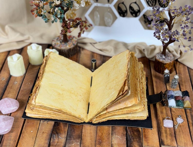 May include: An open, antique-style journal with aged, yellowed pages. The book is bound in black and sits on a wooden surface, surrounded by decorative crystals, candles, and small gemstone trees. The overall aesthetic is mystical and vintage.