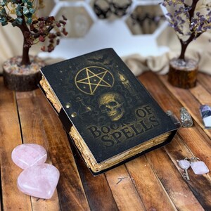 May include: A black "Book of Spells" journal with gold embossed designs, including a pentagram, skull, and candle. The book has aged, yellowed pages and a leather strap closure. Decorative crystals and a small tree are in the background.