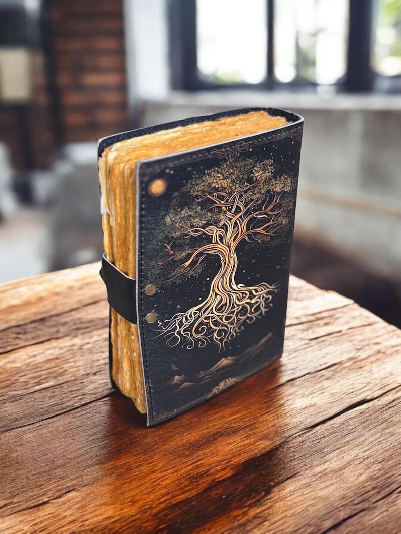 May include: A black leather journal with a gold tree design and a strap closure. The journal has a rustic look and is perfect for writing down thoughts and ideas.