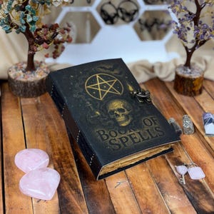 May include: A black book with the words "Book of Spells" in gold lettering. The cover features a gold pentagram, skull, and potion bottle design. The book is secured with a metal clasp and sits on a wooden surface.