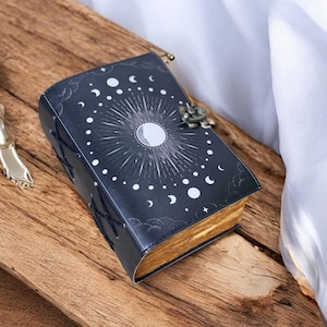 May include: A black leather journal with a celestial design featuring a sun, moon, and stars. The journal has a decorative clasp and is closed.