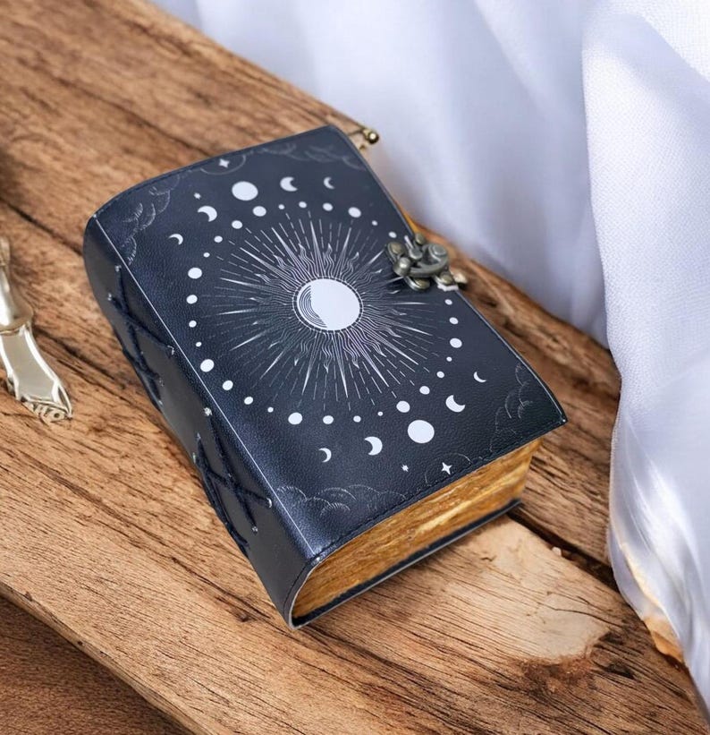 May include: A black leather journal with a celestial design featuring a sun, moon, and stars. The journal has a decorative clasp and is closed.