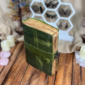 May include: A handmade, green leather journal with a strap closure. The journal has aged, deckled-edge pages and a metal clasp. The cover is a deep green color, and the pages have a warm, aged tone. The journal is closed with a matching green leather strap.
