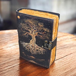 May include: A black leather journal with a gold tree design and a clasp closure. The journal has a distressed look and is perfect for writing or sketching.