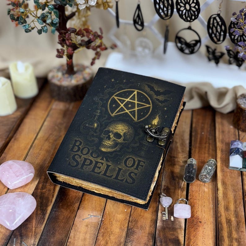 May include: A black leather-bound book with the words "BOOK OF SPELLS" in gold lettering. The cover features a gold pentagram, skull, and potion bottle design. The book is closed with a metal clasp and sits on a wooden surface.