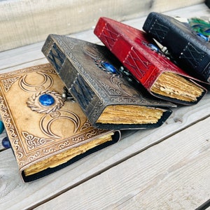 Op de afbeelding: Vier lederen dagboeken met decoratieve omslagen. De dagboeken hebben verschillende gekleurde omslagen: bruin, rood en zwart. Elk dagboek heeft een blauwe edelsteen op de voorzijde. De dagboeken zijn gestapeld op een houten oppervlak.