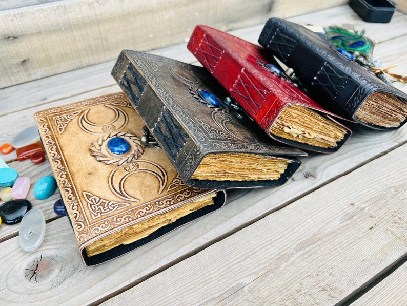 Op de afbeelding: Vier lederen dagboeken met decoratieve omslagen. De dagboeken hebben verschillende gekleurde omslagen: bruin, rood en zwart. Elk dagboek heeft een blauwe edelsteen op de voorzijde. De dagboeken zijn gestapeld op een houten oppervlak.