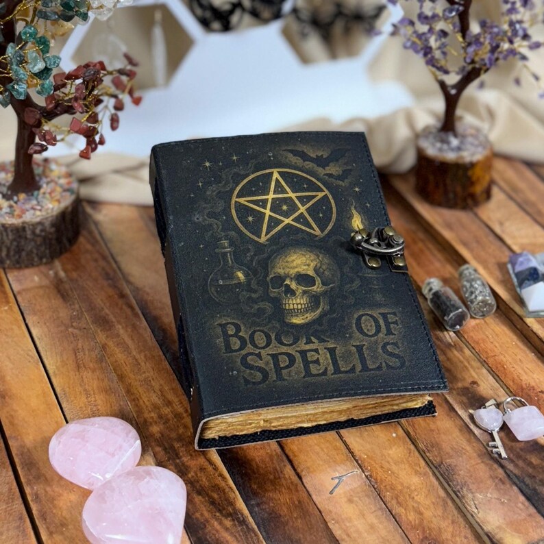 May include: A black leather-bound book with the words "Book of Spells" in gold lettering. The cover features a gold pentagram, skull, and potion bottle design. The book has a metal clasp and aged, yellowed pages.