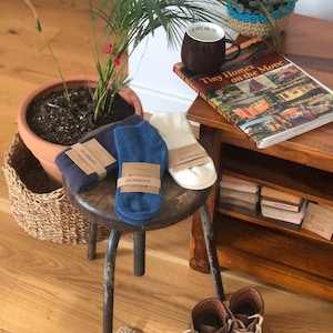 May include: Three pairs of socks in navy, blue, and cream colors, each with a "SACRAMENTO" label. The socks are displayed on a small stool, with a book titled "Tiny Homes on the Move" and a brown mug nearby. Brown leather boots are on the floor.