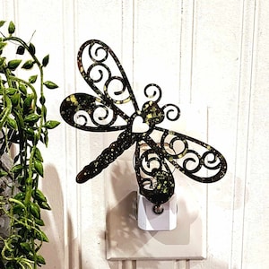 May include: A black metal dragonfly night light plugged into a white electrical outlet. The dragonfly has a decorative design with swirls and is accented with gold glitter.