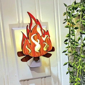 May include: A wooden night light shaped like a campfire with orange and red flames. The night light is plugged into an electrical outlet.