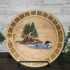 May include: A wooden cribbage board with a loon and trees design. The board has a circular shape and is made of light wood. The design is laser engraved and features a loon swimming in a lake with trees on a small island. The board has a black stand.