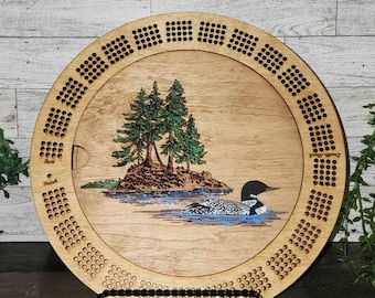 May include: A wooden cribbage board with a loon and trees design. The board has a circular shape and is made of light wood. The design is laser engraved and features a loon swimming in a lake with trees on a small island. The board has a black stand.