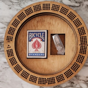 K&ouml;nnte beinhalten: Ein rundes Holzspielbrett mit einem Kartenspiel Bicycle und einem kleinen Beutel mit Murmeln. Das Brett hat eine kreisf&ouml;rmige Bahn mit nummerierten L&ouml;chern und die W&ouml;rter "Start" und "Finish" sind auf dem Brett markiert.