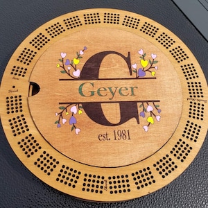 May include: A wooden cribbage board with a personalized wooden inlay featuring the letter "G", the name "Geyer", and the text "est. 1981". The inlay is surrounded by a floral design with pink, purple, and yellow hearts.