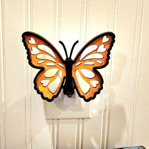 May include: A wooden butterfly night light plugged into an electrical outlet. The butterfly is orange with black accents and has a cutout design.
