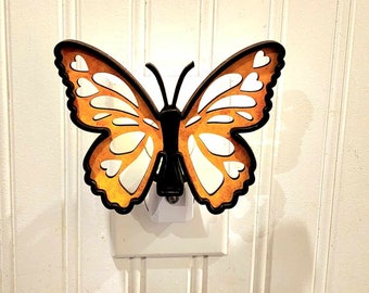 Könnte beinhalten: Ein hölzernes Schmetterlings-Nachtlicht, das in eine Steckdose gesteckt wird. Der Schmetterling ist orange mit schwarzen Akzenten und hat ein ausgeschnittenes Design.