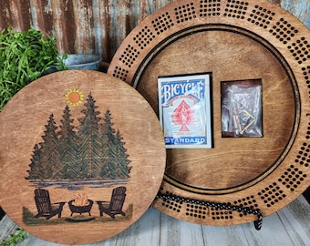May include: A wooden game board with a black string around the edge. The board has a circular design with a central area that is a lighter wood color. The central area has a playing card and a small box with a metal object inside. The other side of the board has a design of a campfire with trees and a lake.