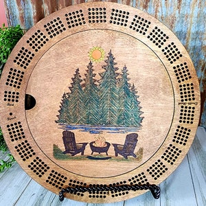 K&ouml;nnte beinhalten: Ein rundes Holzspielbrett mit einer Camping-Szene mit einem Lagerfeuer, zwei St&uuml;hlen und einem See. Das Brett hat einen schwarzen und wei&szlig;en Gitterrand. Der Text "Start" und "Finish" ist auf dem Brett zu sehen.