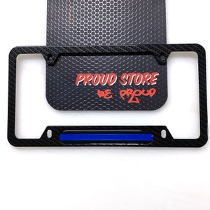 May include: Black carbon fiber license plate frame with a blue thin blue line stripe across the bottom. The frame has two holes on each side for mounting. The text "PROUD STORE BE PROUD" is visible in the background.