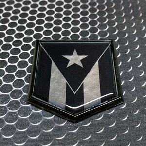 K&ouml;nnte beinhalten: Ein schwarzes und silbernes schildf&ouml;rmiges Emblem mit einem wei&szlig;en Stern und drei Streifen. Das Emblem ist aus gl&auml;nzendem Material gefertigt und hat einen schwarzen Rand.
