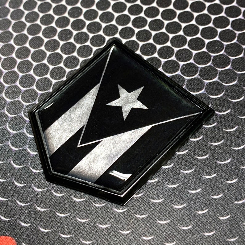 K&ouml;nnte beinhalten: Ein schwarzer und silberner schildf&ouml;rmiger Aufkleber mit der Flagge von Puerto Rico. Die Flagge zeigt einen wei&szlig;en f&uuml;nfzackigen Stern auf einem roten Dreieck, mit drei wei&szlig;en Streifen auf schwarzem Hintergrund.