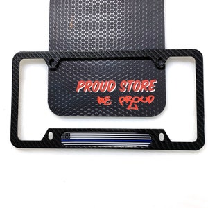 May include: Black carbon fiber license plate frame with a thin blue line American flag design. The frame has two holes on each side for mounting. The text "PROUD STORE BE PROUD" is printed on the background.