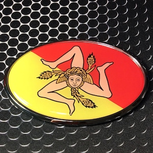 May include: A chrome-plated oval car emblem with a yellow and red background. The emblem features a stylized image of a woman with three legs, a head with a wreath, and wheat stalks.