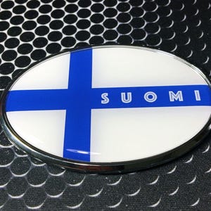 May include: Oval emblem featuring the Finnish flag design. The emblem has a white background with a blue cross and the word "SUOMI" in white letters. The emblem is framed with a silver border.