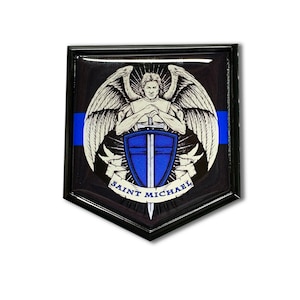 May include: A black and blue enamel pin with a depiction of Saint Michael, an angel with wings, holding a sword and shield. The pin has a thin blue line across the top and the words "SAINT MICHAEL" at the bottom.