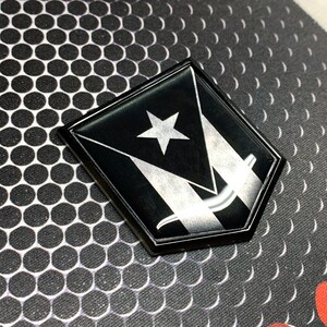 K&ouml;nnte beinhalten: Ein schwarzes und silbernes Metallemblem mit einem wei&szlig;en Stern und einem stilisierten Buchstaben "M" auf einem schwarz-wei&szlig;en Wabenhintergrund.