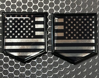 May include: Two black and silver American flag emblems with a thin blue line. The emblems are in a hexagon shape and have a glossy finish.