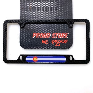 May include: Black carbon fiber license plate frame with a Colorado state flag design. The frame has two holes on the top and two holes on the bottom for mounting. The text "PROUD STORE BE PROUD" is visible on the background.