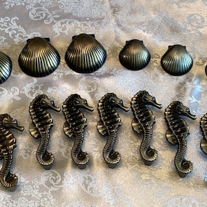 Könnte beinhalten: Ein Satz von acht antiken goldfarbenen Schubladenknöpfen in Form von Seepferdchen und Muscheln. Die Seepferdchenknöpfe sind detailliert und haben einen gekräuselten Schwanz. Die Muschelknöpfe sind glatt und haben eine gerippte Textur.