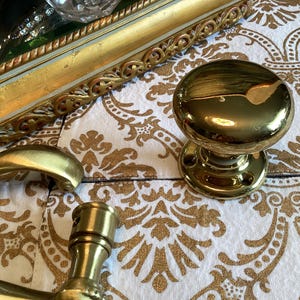May include: A polished brass door knob with a smooth, rounded surface and a circular mounting plate featuring two screw holes. The knob has a reflective, metallic finish and is positioned alongside a brass door handle and a decorative gold frame.