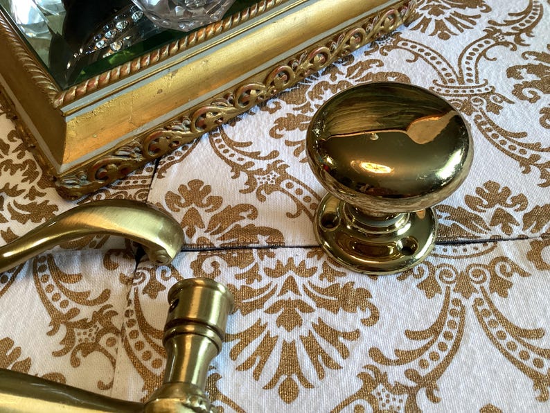 May include: A polished brass door knob with a smooth, rounded surface and a circular mounting plate featuring two screw holes. The knob has a reflective, metallic finish and is positioned alongside a brass door handle and a decorative gold frame.