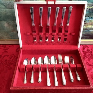 May include: A silver flatware set displayed in a red velvet-lined case. The set includes knives, spoons, forks, and serving pieces. The case is open, revealing the neatly arranged silverware. The case has a white exterior and is set on a red patterned tablecloth.