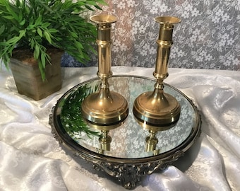 May include: Two brass candlesticks with a classic design, displayed on a mirrored tray with an ornate silver-colored edge. The candlesticks have a cylindrical body and a flared top. The background includes a potted plant and a white floral patterned fabric.
