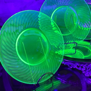 May include: A collection of glowing green glass plates. The plates have a circular shape with a textured, swirled design. The plates are displayed on a decorative stand, and the scene is illuminated with a purple light.