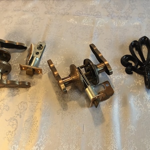 Può includere: Un set di tre maniglie per porte in ottone antico e un gancio decorativo in metallo nero. Le maniglie delle porte sono in diversi stati di smontaggio, mostrando i meccanismi interni. Il gancio ha un design floreale.